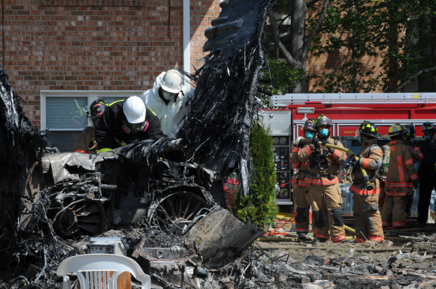 Feuerwehrleute in Schutzausrüstung löschen ein Gebäude Feuer, mit verkohlten Überresten einschließlich eines Fahrzeugs, Stuhl und Pflanzen sichtbar auf dem Boden.