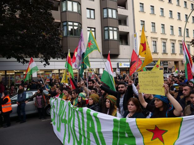Eine große Gruppe von Menschen marschiert mit Flaggen und Schildern die Straße entlang, mit einem geparkten Auto auf der rechten Seite und einem Baum auf der linken Seite, vor einem Hintergrund von Gebäuden mit Fenstern und Namensschildern, was auf eine algerische Demonstration hinweist.