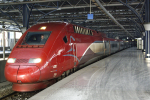 Ein roter Hochgeschwindigkeitszug steht auf einem Bahnhof mit einer Plattform auf der rechten Seite, einige Menschen stehen und Säulen und Stangen im Hintergrund.