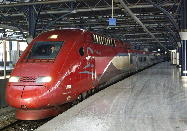Ein roter Hochgeschwindigkeitszug steht auf einem Bahnhof mit einer Plattform auf der rechten Seite, einige Menschen stehen und Säulen und Stangen im Hintergrund.