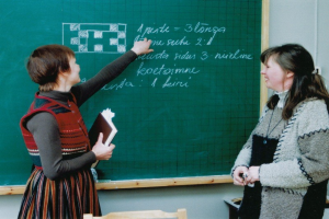 Zwei Frauen in einem Klassenzimmer, eine hält ein Buch und die andere schreibt an einer Tafel, mit Stühlen unten sichtbar.