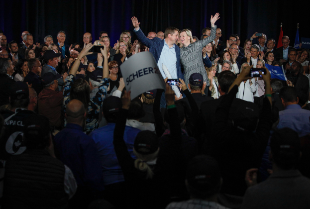 Ein Mann und eine Frau stehen vor einem Publikum, einige halten Schilder und Handys, im Hintergrund sind Vorhänge und Fahnen zu sehen.