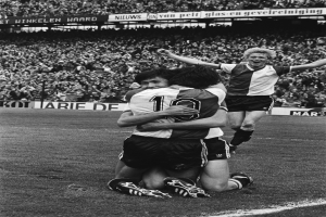 Drei Fußballspieler feiern ein Tor auf dem Boden in einem schwarz-weißen Stadion vor einer jubelnden Menge.