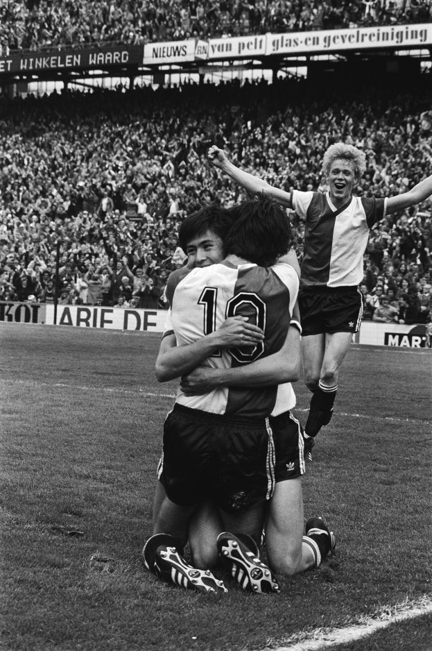 Drei Fußballspieler feiern ein Tor auf dem Boden in einem schwarz-weißen Stadion vor einer jubelnden Menge.