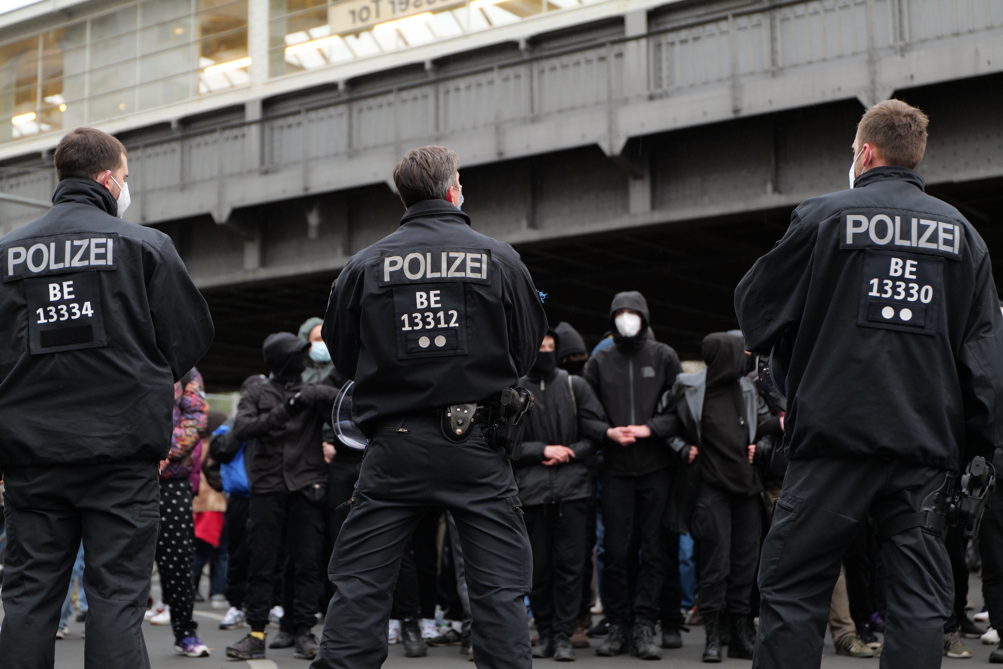 Police officers in black uniforms and masks stand in front of a crowd during a protest, with a bridge and building visible in the background.