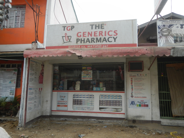 Ein Apotheken-Schild mit der Aufschrift "The Generics Pharmacy" ist an einem Gebäude in einer städtischen Gegend mit sichtbarer Infrastruktur und Fahrzeugen unter einem klaren Himmel zu sehen.
