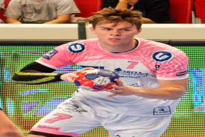 Ein Mann in einem pinken Hemd hält einen Fußball, wobei die Hand einer anderen Person links zu sehen ist; im Hintergrund sitzen ein paar Menschen auf Stühlen, was auf ein Handballspiel hindeutet.