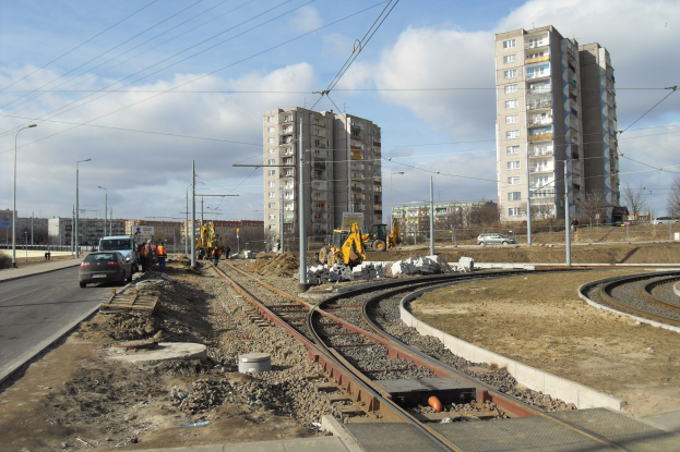 Baustelle mit einer durchlaufenden Bahnschiene, umgeben von Geb├Ąuden, Stra├čeninfrastruktur, Kraftfahrzeugen, Passanten, verstreuten Steinen, B├Ąumen und einem bew├Âlktem Himmel.