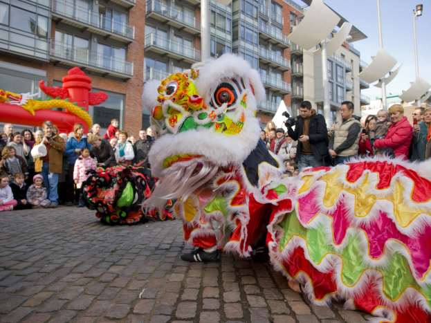 Ein lebendiges chinesisches Neujahrsfest in Amsterdam mit einer Löwen-Tanzvorstellung vor einer Zuschauermenge, darunter einige, die das Ereignis fotografieren, vor Buildings und einem klaren blauen Himmel.