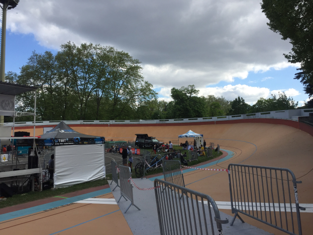 Eine Gruppe von Radfahrern auf einer Schotterstrecke bei den UCI BMX-Weltmeisterschaften 2018, mit Zelten, Fahrzeugen, Geländern, Bäumen und einem bewölkten Himmel im Hintergrund.