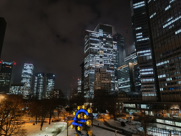 Europäische Zentralbank-Hauptquartier in Frankfurt, Deutschland bei Nacht, mit beleuchteten Gebäuden, Bäumen, Straßenlaternen, Lichtern und Fußgängern unter einem bewölkten Himmel, mit Wasserzeichen.