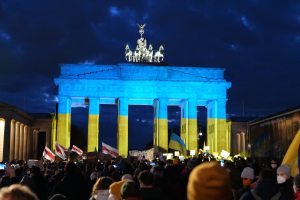 Menschenmenge mit Fahnen und Schildern vor dem Brandenburger Tor in Berlin, mit einer Fahne auf der rechten Seite des Bildes.