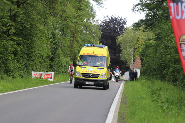Ambulanz fährt auf einer Straße mit Radfahrern nebenher, umgeben von Gras, Bäumen, Häusern, Strommasten und einem klaren blauen Himmel.