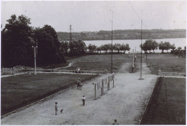 Ein Schwarz-Weiß-Foto von Menschen, die Fußball spielen, umgeben von Bäumen, Pfählen und einem Zaun, mit einem Gewässer und einem klaren Himmel im Hintergrund und Text am unteren Rand des Bildes.