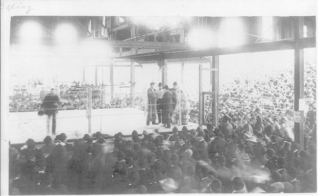Schwarz-weiß-Foto einer Menge vor einer Bühne mit Menschen darauf und im Publikum, was auf einen Boxkampf in einem Stadion hinweist.