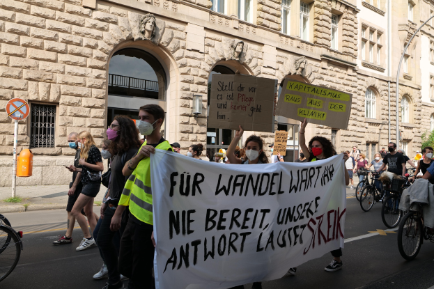 Eine Gruppe von Menschen, die auf einer Straße gehen, einige halten Schilder und Banner, andere fahren Fahrräder, mit einem Gebäude im Hintergrund, das Fenster, Bögen, Säulen und Skulpturen hat, Bäume und eine Straßenlaterne, nehmen an einer Protestaktion gegen die Anti-Kriegs-Bewegung in Berlin teil.