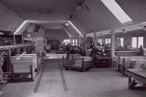 Ein Schwarz-Weiß-Foto eines Fabrikinnenraums mit Arbeitern, Tischen, mit Holz gefüllten Transportwagen und Deckenlampen, dessen Wände mit Fenstern gesäumt sind.