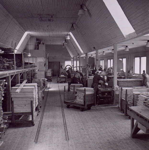 Ein Schwarz-Weiß-Foto eines Fabrikinnenraums mit Arbeitern, Tischen, mit Holz gefüllten Transportwagen und Deckenlampen, dessen Wände mit Fenstern gesäumt sind.