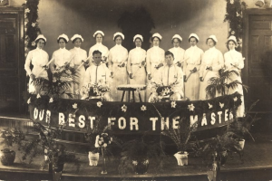 Gruppe von Frauen in weißen Kleidern und Häubchen auf einer Bühne mit einem zentralen Banner, auf dem 'Unsere Besten für den Meister' steht, umgeben von Blumentöpfen und vor Türen und einer Wand