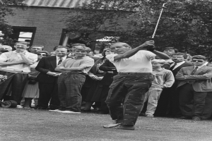 Ein Mann schwingt einen Golfschläger vor einer Menge, mit einem Gebäude und Bäumen im Hintergrund, dargestellt in Schwarz-Weiß.