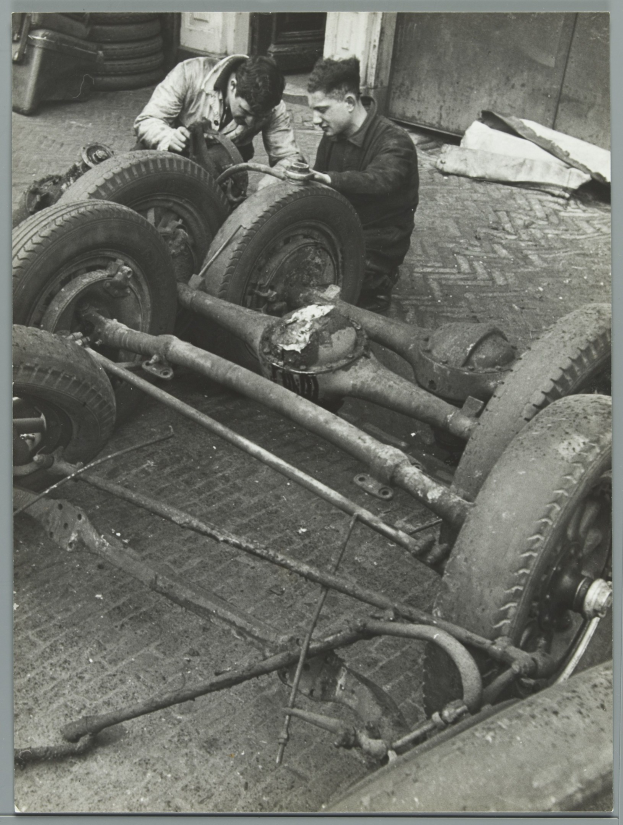 Zwei Männer in einer schwarz-weißen Garage reparieren ein Auto und halten Werkzeuge, mit Reifen und anderen Gegenständen in der Nähe.