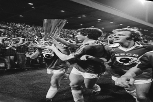 Eine Gruppe von Männern, die einen Pokal auf einem Fußballfeld halten und ihren Sieg im FA Cup Final feiern, umgeben von einer Menge hinter einem Zaun, unter Stadionbeleuchtung, in Schwarz-Weiß.