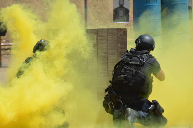 Zwei Polizeibeamte in Einsatzausrüstung mit gelbem Rauch aus dem Mund, stehend in der Nähe einer Wand mit Text und anderen Gegenständen im Hintergrund.
