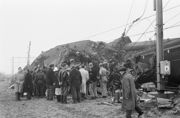 Schwarz-weißes Bild einer Gruppe von Menschen in Mützen, die neben einem Zug stehen, der gegen einen Pfahl gekracht ist, mit Strommasten, Drähten, Bäumen und einem klaren Himmel im Hintergrund.