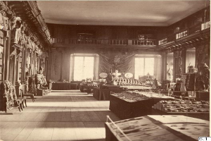 Ein altes Schwarz-Weiß-Foto eines Raums voller Tische, Stühle, Skulpturen an den Wänden, Fenstern, Vorhängen, Geländern und anderen Gegenständen mit der Aufschrift "The Museum of Fine Arts, London" unten.