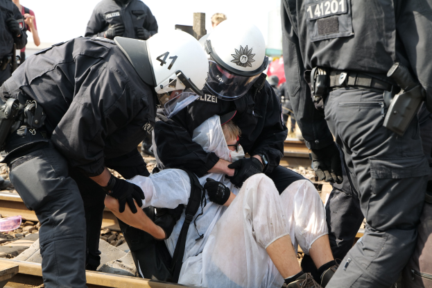 Eine Gruppe von Polizisten in Schutzausrüstung verhaftet einen Mann in einem weißen Kleid auf einem Bahngleis, während andere im Hintergrund stehen.