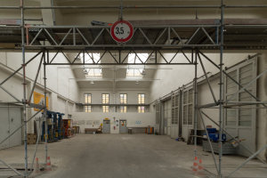 Großer Lagerraum mit ausgedehnten Gerüsten, Verkehrskegeln, Metallstangen, einem Schild, Maschinen, einer Tür, Fenstern, Wänden, Deckenleuchten und Dach.