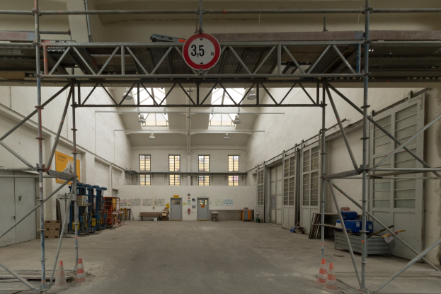 Großer Lagerraum mit ausgedehnten Gerüsten, Verkehrskegeln, Metallstangen, einem Schild, Maschinen, einer Tür, Fenstern, Wänden, Deckenleuchten und Dach.