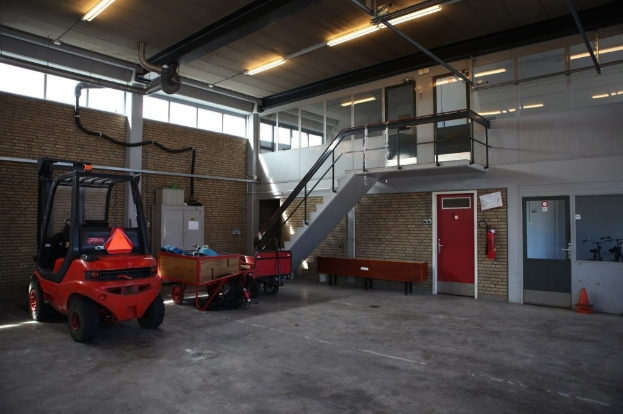 Großer Lagerraum mit einem roten Gabelstapler, einem Wagen mit Gegenständen, einer Bank, einem Feuerlöscher, einer Treppe mit Geländer, Türen, Fenstern, Rohren und einem sichtbaren Rad.