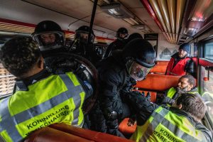 Polizeibeamte in Einsatzausrüstung auf einem Bus sitzend, mit einer Person in der Mitte, sichtbaren Busfenstern und einem Plakat an der Wand im Hintergrund.
