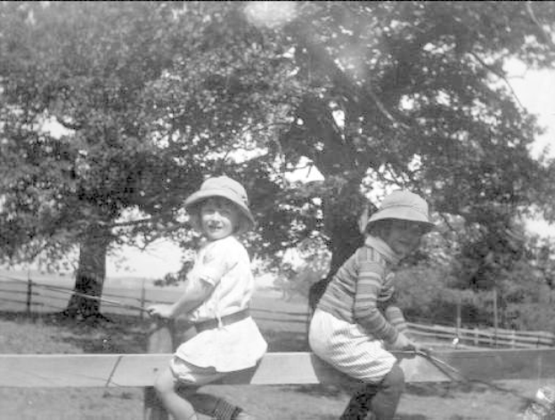 Zwei junge Jungs in Hüten mit Baseballschlägern auf einem grasigen Park mit Bäumen und einem Zaun im Hintergrund, Schwarz-Weiß-Foto.