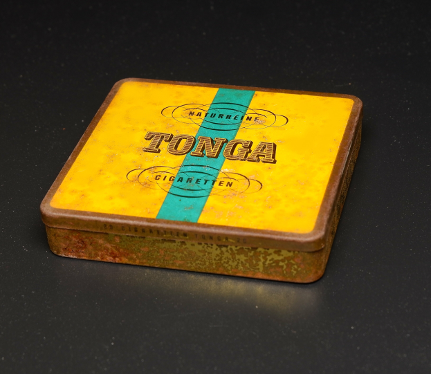 Eine gelbe und blaue Dose Tonga Zigaretten auf einem schwarzen Tisch, mit Text, der die Marke und Art der Zigaretten angibt.