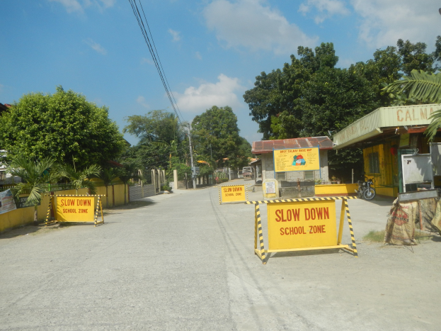 Gelbes "Langsam Schulzone"-Schild auf der linken Seite einer Straße mit geparkten Fahrzeugen auf der rechten Seite, Bäumen, Gebäuden, Strommasten, Drähten und Himmel im Hintergrund.