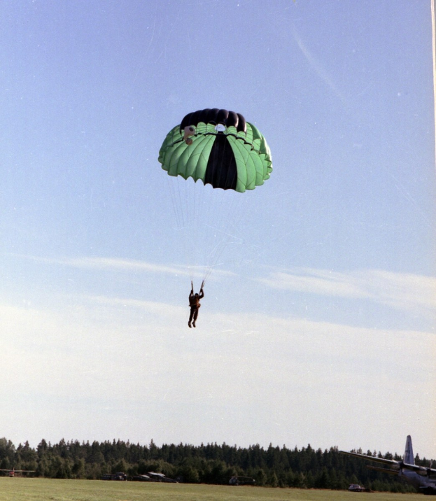 Eine Person beim Paragleiten in der Luft mit einem Fallschirm, umgeben von Gras und Bäumen, mit einem Flugzeug rechts im Bild und Wolken am Himmel.