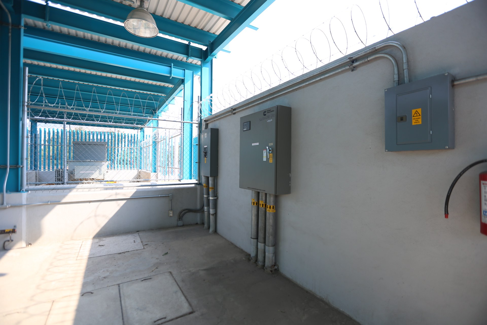 Ein Raum in einer elektrischen Umspannstation mit einem Feuerlöscher auf der rechten Seite, elektrischen Kästen und Rohren an der Wand, einer Deckenleuchte und einem Zaun im Hintergrund.