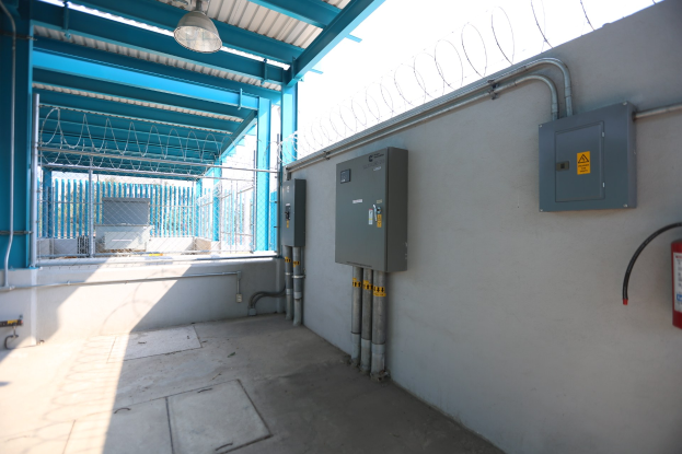 Ein Raum in einer elektrischen Umspannstation mit einem Feuerlöscher auf der rechten Seite, elektrischen Kästen und Rohren an der Wand, einer Deckenleuchte und einem Zaun im Hintergrund.