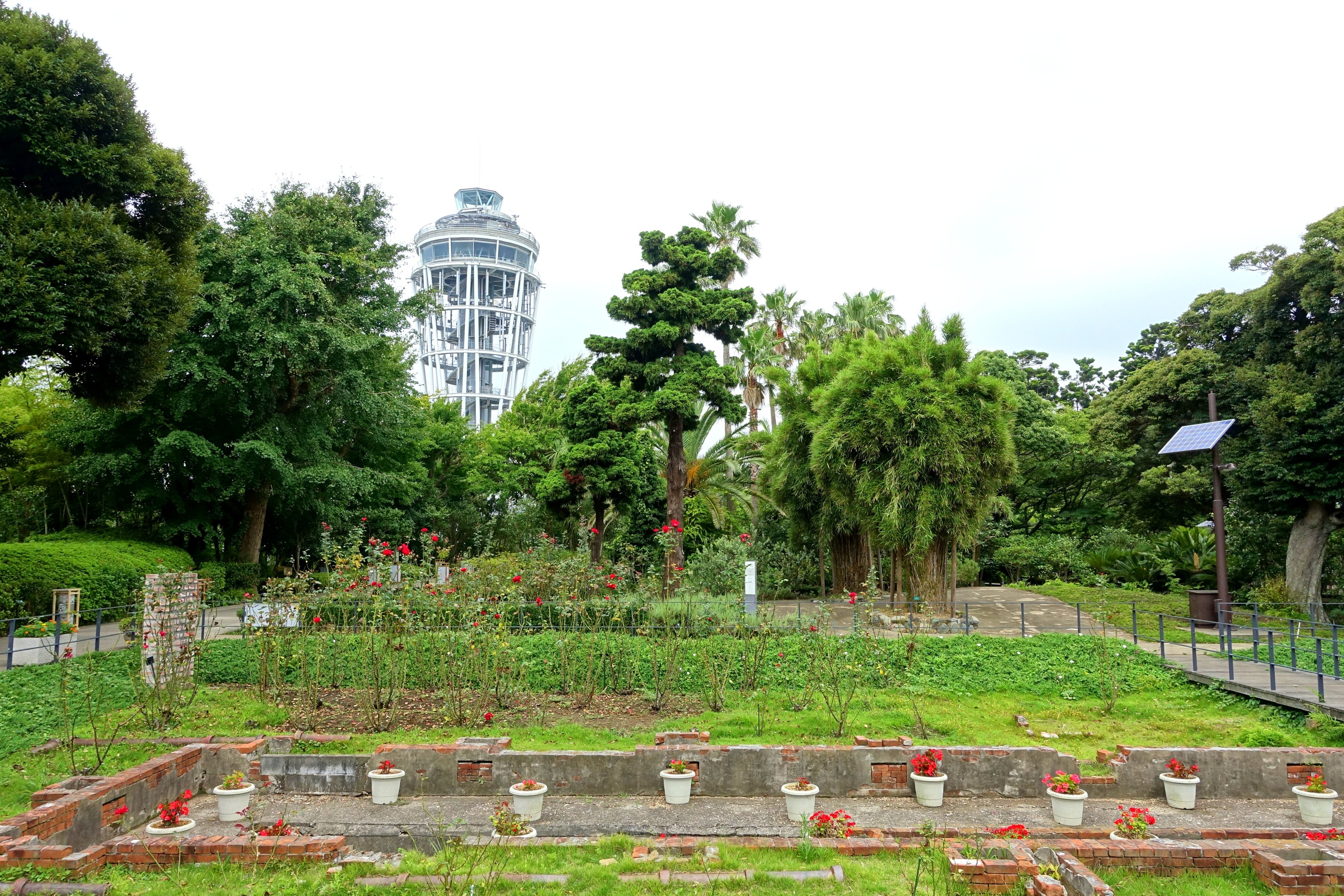 Ein Park mit einem Turm im Hintergrund, umgeben von grünem Gras, Pflanzen, Bäumen und verstreuten Blumentöpfen, mit einer Straße mit Geländern und einer Pfostenmontage unter einem sichtbaren Himmel.