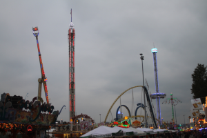 Eine Menschenmenge versammelt sich um einen hell erleuchteten Faschingsumzug in der Nacht, mit beleuchteten Fahrgeschäften, Pfählen und Bäumen unter einem bewölkten Himmel.