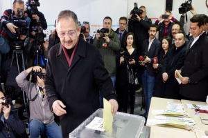 Ein Mann in einem schwarzen Mantel und Brille stimmt in einer Wahllokal umringt von Menschen mit Kameras und Mikrofonen, Tischen mit Papieren und Gegenständen und einer Wand mit Uhr und Lautsprecher ab.