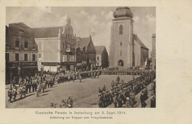 Ein Schwarz-Weiß-Foto eines Umzugs in Insterburg am 5. September 1914 mit vielen Menschen, Gebäuden im Hintergrund und Text am unteren Rand des Bildes.