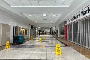 Das Innere eines Einkaufszentrums mit Bodenaufschriften, die aufgrund der Covid-19-Pandemie auf den Schließung hinweisen, eine getextete Wand auf der rechten Seite, Gegenstände auf der linken Seite, Deckenleuchten und einige Personen im Hintergrund.