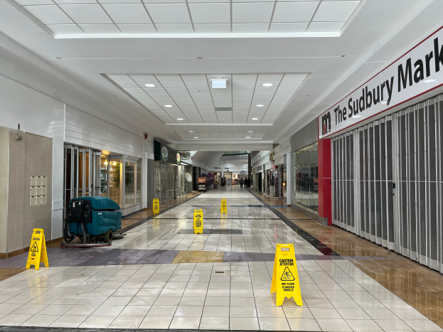 Das Innere eines Einkaufszentrums mit Bodenaufschriften, die aufgrund der Covid-19-Pandemie auf den Schließung hinweisen, eine getextete Wand auf der rechten Seite, Gegenstände auf der linken Seite, Deckenleuchten und einige Personen im Hintergrund.