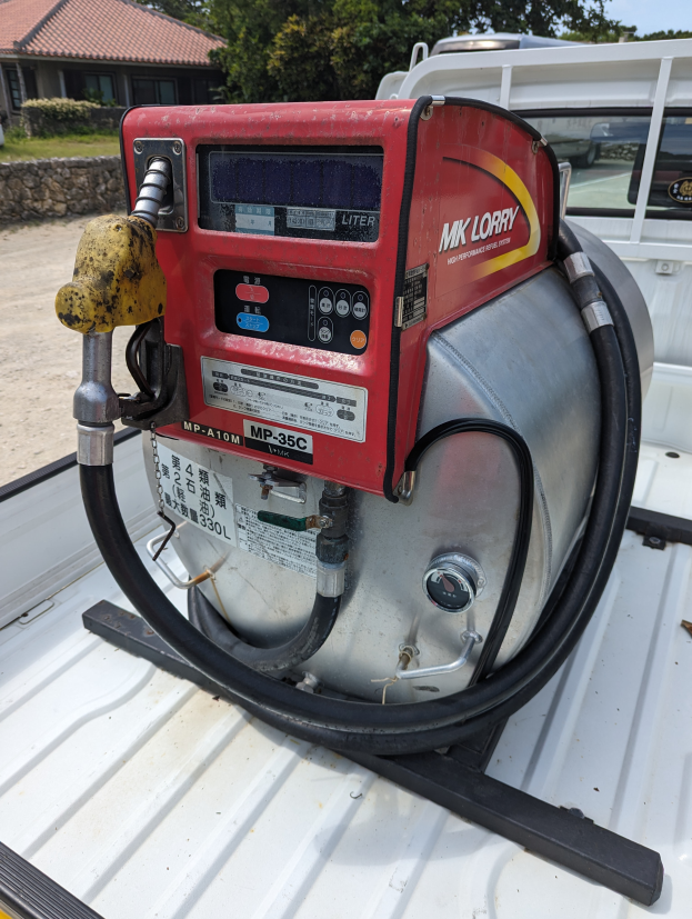 Ein Lastwagen mit einer Tankstelle auf der Ladefläche, umgeben von Bäumen, einem Haus und einem klaren blauen Himmel, ausgestattet mit einem Motor und einer Kraftstoffabgabe für die Betankung von Fahrzeugen.
