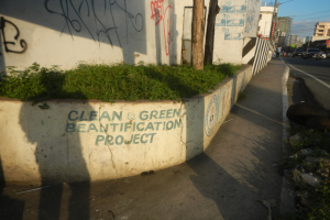 Eine Straße mit Fahrzeugen, Gebäuden, Bäumen, Pfählen, Drähten und einer Wand mit Graffiti 'Saubere und grüne Verschönerungsprojekt' mit Pflanzen im Vordergrund und einem bewölkten Himmel im Hintergrund.