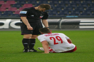 Ein Fußballspieler sitzt neben einem Schiedsrichter auf dem Boden, beide in Sportkleidung, mit Stadionplakaten und Stühlen im Hintergrund.