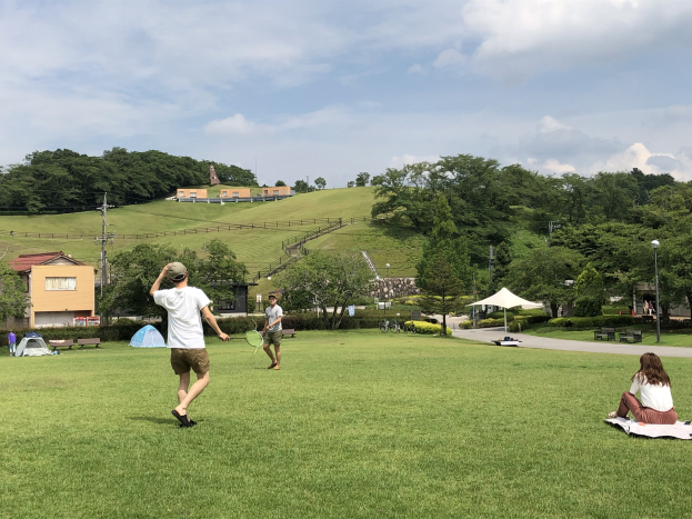 Eine Gruppe von Menschen, die Badminton in einem Park spielt, mit Zelten, Straßenlaternen und Gebäuden im Hintergrund.